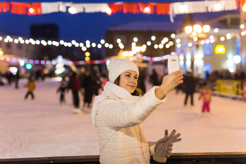 Obraz premium Christmas, winter, technology and leisure concept - happy young woman taking picture with smartphone on ice skating rink outdoors.