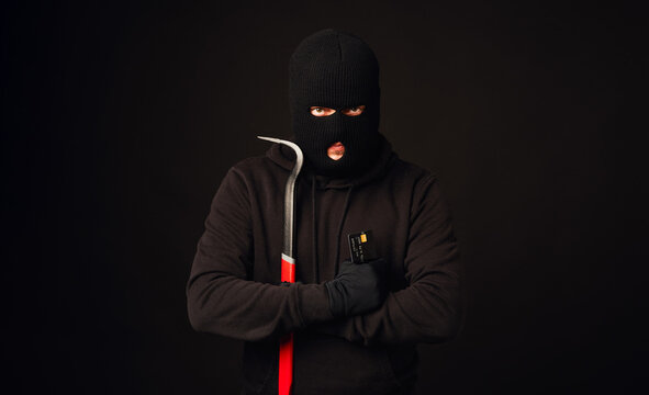 Thief in black balaclava holding crowbar and black credit card. Banking and non cash thief concept