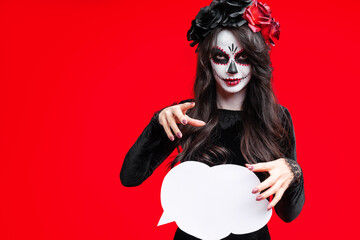 Girl with creative sugar skull makeup in wreath of flowers on head, isolated on red background hold white cloud. Holiday concept Dia De Los Muertos poster for Halloween party or La Calavera Catrina.