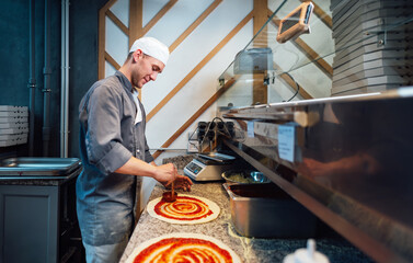 A glad chef spreads the sauce on the pizza dough. Catering kitchen work.