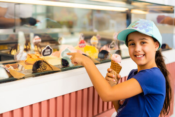 A little girl is standing near a shop window with an assortment of ice-cream and sweets. The child...