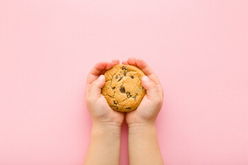 Baby girl hands holding dry cookie with chocolate pieces on light pink table background. Pastel color. Closeup. Point of view shot. Sweet snack. Top down view.
