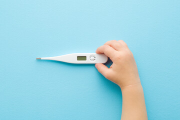 Baby hand holding white digital thermometer on light blue table background. Pastel color. Closeup....