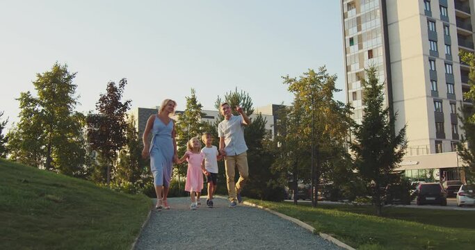 The family is walking in the park on a cloudless day. Parents hold their children by the hand and walk along the path. Around them there is green grass, trees. You can see houses from the side
