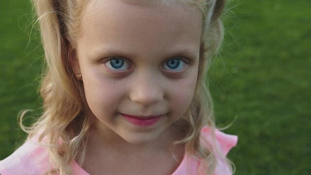 A little girl in a pink dress and blue eyes looks into the camera close-up. She has blonde wavy hair. She smiles shyly