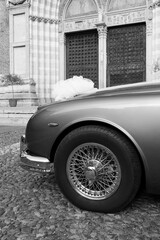 Part of a beautiful wedding car with a front wheel and a bow on the hood against the background of an old church. Vertical black and white photo