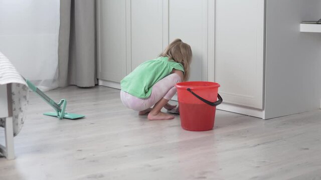A little girl washes the floors of the house with a sex rag, the baby washes a dirty rag in a bucket of water.