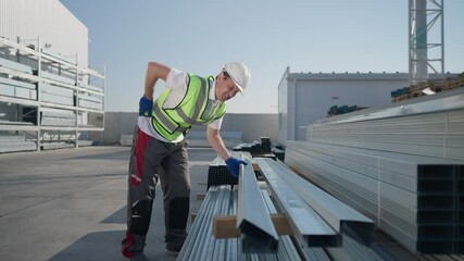 Wide shot Caucasian man having backache lifting heavy manufactured cargo at warehouse outdoors. Sad unwell worker in hard hat holding back sighing with painful facial expression. Slow motion