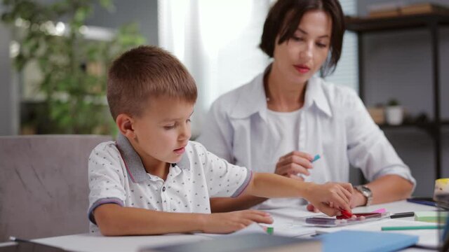 Caucasian woman with dark hair sitting with cut boy at desk and drawing in album with colorful pencils. Mother with son spending time at home usefully.
