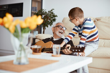 Happy grandfather and grandson have fun while playing acoustic guitar at home.