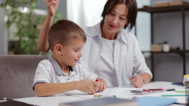 Happy boy and his charming mother sitting together at desk and drawing with colorful pencils in album. Creative process at home. Education moment during parenthood.