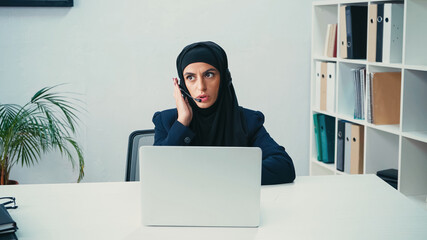 Muslim woman in headset using microphone and talking near laptop in call center