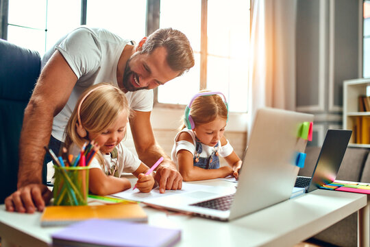 Online Training. Two Schoolgirl Sisters In Headphones Listen To Lessons On Laptops. Dad Helps Daughters Study. School At Home In A Pandemic And Quarantine.