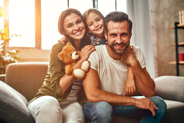 Happy dad and mom with their cute daughter and teddy bear hug and have fun sitting on the sofa in...