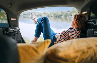 Woman dressed in warm calm colors clothes and jeans lying in the cozy car trunk and enjoying the...