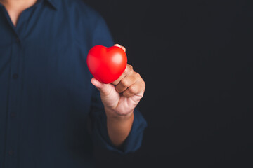 A young man in a blue shirt holding a red heart shape while standing with a black background in the studio. Close-up photo. Space for text. Donate, love, and encouragement concept
