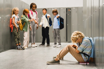 Sad crying schoolboy pupil sitting on the floor at the school hall while his classmates teenagers...