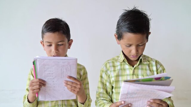 close shot of two little adorable primary school children wearing uniforms holding books in their hands and reading against a white background or a wall. learning and education concept