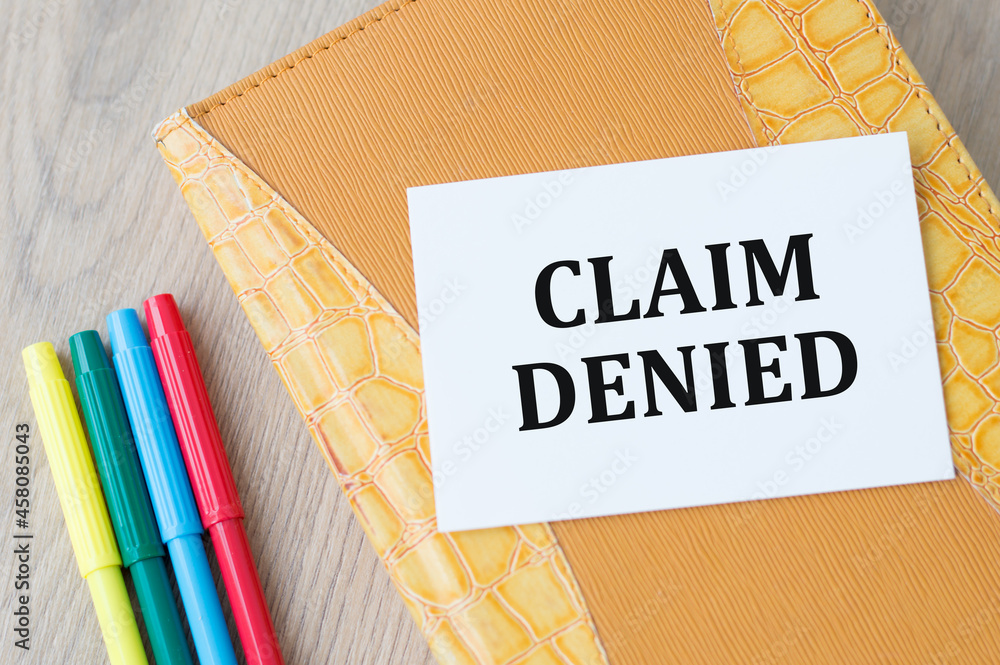 Poster card on a notepad with the text claim denied, next to colored markers on a wooden background