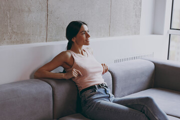 Side view profile excited vivid young latin woman 20s in casual clothes sit on sofa enjoy free time rest relax spend time in living room in own house be lost in reverie good day. Real estate concept.
