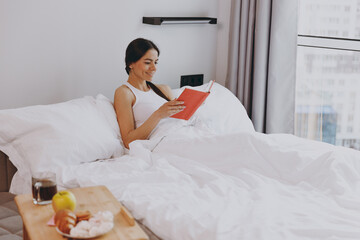 Obraz na płótnie Canvas Happy smiling vivid young woman in white tank top sit in bed reading book rest relax spend time in bedroom lounge home in own room house wake up dream be lost in reverie good day. Real estate concept.
