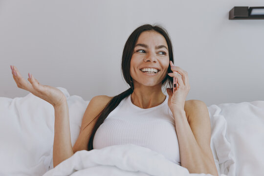 Happy young woman in white tank top sit in bed talk speak on mobile cell phone conducting pleasant conversation look aside in bedroom lounge home in own room house wake up be lost in reverie good day.