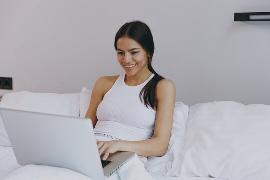 Excited beautiful fun young woman in white tank top sit in bed use work on laptop pc computer chtting typing browsing in bedroom lounge home own room house wake up dream be lost in reverie good day