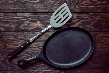 Empty cast iron frying pan and a culinary spatula on wooden background, top view