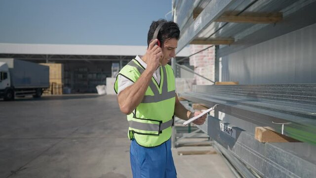 Side view of cheerful expert Middle Eastern man dancing to music in headphones checking bill of lading and delivered goods at warehouse outdoors on sunny day. Positive confident employee working