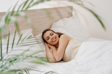 Smiling young woman in casual casual clothes lying in bed hands folded under head look camera rest...