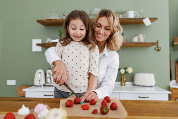 Happy scared chef baker mom woman in white shirt work with child baby girl helper teach to cook cut together at kitchen table home . Cooking food process concept Mommy little kid prepare fruit cake