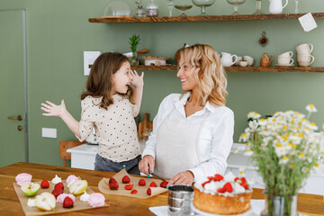 Happy chef cook baker mom woman in white shirt work with child baby girl helper teach baking spread arms at kitchen table home Cooking food process concept Mommy little kid daughter prepare fruit cake