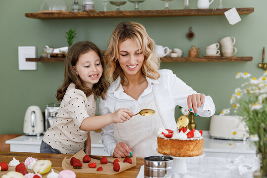 Happy fun chef cook baker mom woman in white shirt work with child baby girl helper decorate pie with cream at kitchen table home. Cooking food concept Mommy little kid daughter prepare fruit cake