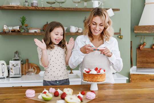 Happy chef cook baker mom woman in white shirt work with baby girl helper take photo on mobile cell phone at kitchen table home. Cooking food process concept Mommy little kid daughter prepare cake
