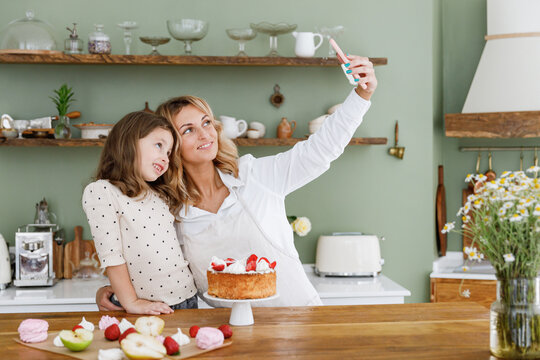 Happy chef cook baker mom woman in white shirt work with baby girl helper do selfie shot on mobile cell phone at kitchen table home Cooking food process concept Mommy little kid daughter prepare cake