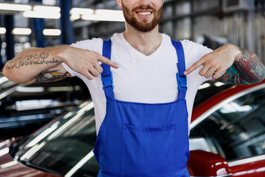 Close up cropped happy young male technician car mechanic man wears denim blue overalls white t-shirt point on himself work in light modern vehicle repair shop workshop indoors Tattoo traslate fun.