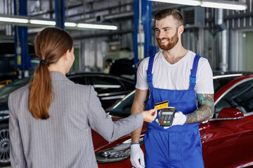 Young happy cheerful professional car mechanic man in blue overalls gloves hold payment terminal fow paying with credit card by female owner driver woman work in vehicle repair shop workshop indoors - obrazy, fototapety, plakaty