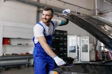 Strong smiling troubleshooter young male professional technician car mechanic man in denim blue overalls white t-shirt raised hood look aside work in light modern vehicle repair shop workshop indoor.