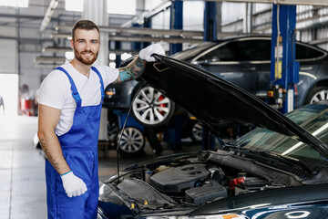 Fototapeta na wymiar Happy troubleshooter young male professional technician car mechanic man in denim blue overalls white t-shirt fixing problem with raised hood bonnet work in modern vehicle repair shop workshop indoor.