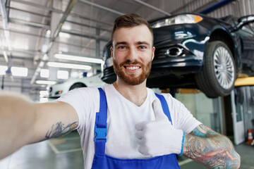 Close up fun young professional technician mechanic man in blue overalls t-shirt do selfie shot pov mobile phone stand near car lift show thumb up gesture work in vehicle repair shop workshop indoors.