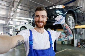 Close up fun young professional technician mechanic man in blue overalls t-shirt do selfie shot pov mobile phone stand near car lift point thumb on himself work in vehicle repair shop workshop indoor