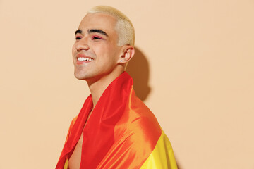 Side view fun young cheerful blond latin gay man with make up in beige tank shirt wrapped in rainbow flag looking aside isolated on plain light ocher background studio People lgbt lifestyle concept.