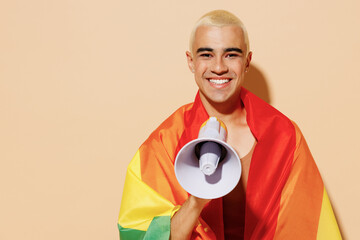 Young happy latin gay man with make up in beige tank shirt wrapped in rainbow flag hold shout aside in megaphone isolated on plain light ocher background studio portrait People lgbt lifestyle concept.