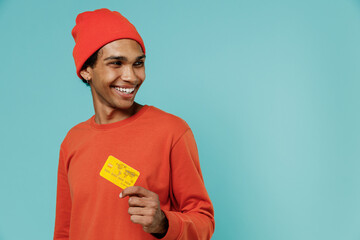 Young smiling happy cool african american man 20s in orange shirt hat hold in hand credit bank card...