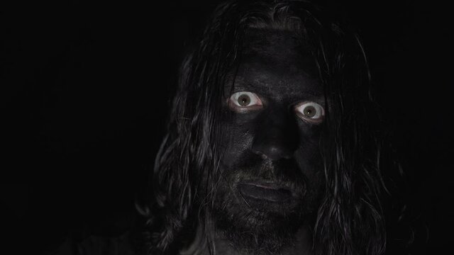 A scary man with black painted face. Opening his eyes and staring into the camera. Low light and dark background. Horror concept.