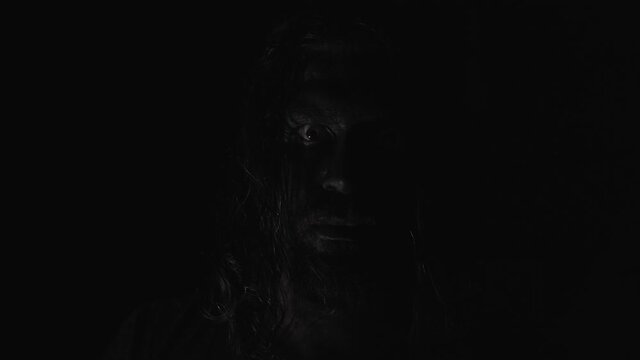 A scary man with black painted face staring into the camera. The light moving from left to right around his face. Low light and dark background. Horror concept.