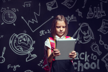 Fototapeta na wymiar Cute schoolgirl preparing to go to school with backpack looking at the tablet. Back to school
