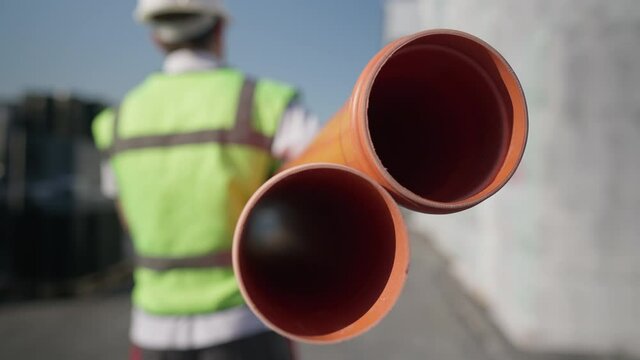 Live camera follows orange plastic pipes with blurred man in uniform carrying warehouse product. Back view of unrecognizable employee walking in slow motion with cargo for shipping outdoors