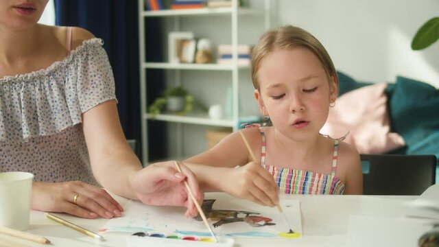 Young mother teaching her daughter to draw. Lovely babysitter drawing with little girl at home, using watercolor. Motherhood and happy childhood concept. Preschool children education.