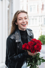 young happy woman in leather jacket holding red roses on urban street in europe